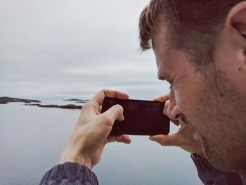 Side view of mid adult man photographing sea with smart phone while standing against cloudy sky