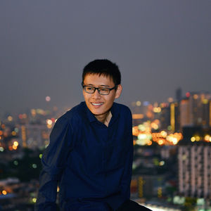 Young man sitting against illuminated city at dusk