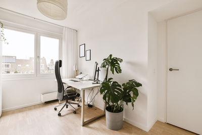 Office chair placed at wooden table with computer screen in spacious white room in flat designed in minimal style