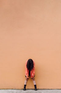 Full body of young anonymous female covering face with long brown hair bending forward while standing against orange wall