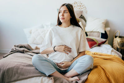 Pregnant caucasian brunette  mom sitting on the bed in the bedroom holding her pregnant tummy.