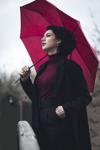 Woman looking away holding umbrella standing against sky