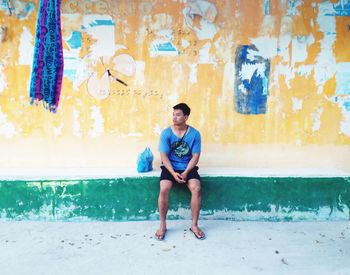 Full length portrait of young man sitting against wall