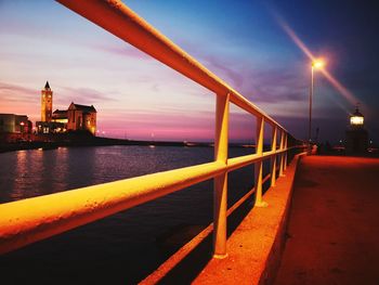 Illuminated street by sea against sky at sunset