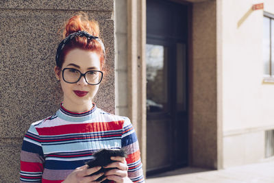 Portrait of young female hipster holding mobile phone while standing against wall