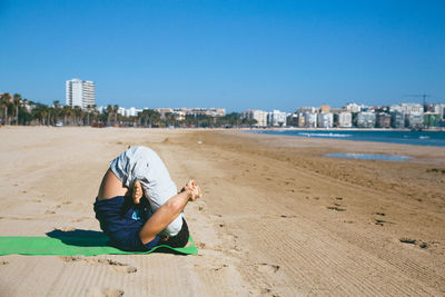 Full length of man performing yoga at beach by city against clear blue sky