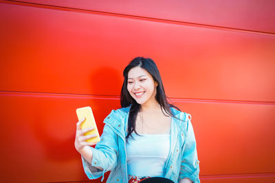 Smiling young woman taking selfie from mobile phone by red wall