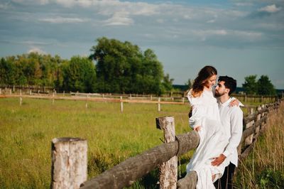 Happy wedding couple by fence on field against sky