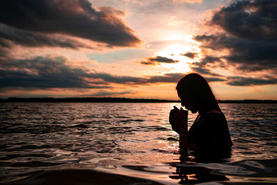 A young woman drinking from a mug while sitting in a lake at sunset 