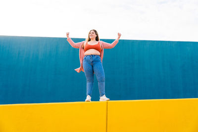 Full length of young woman with arms raised standing against wall