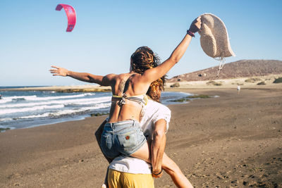 Rear view of man piggybacking woman at beach against clear sky