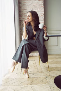 Ballerina in a suit with a cup of coffee smiling and dreaming is sitting at a table by the window