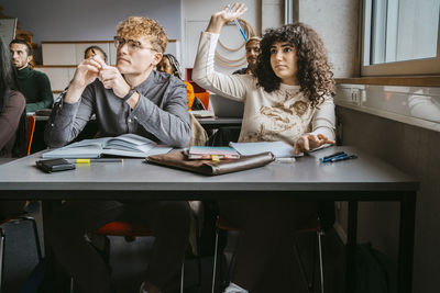 Young female student sitting with hand raised by friends in classroom