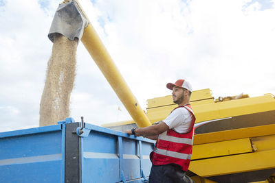 Side view of male farmer standing near industrial combine harvester unloading dried grains into trailer while working in agricultural plantation