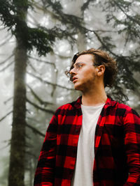 Young man looking away while standing against trees in forest