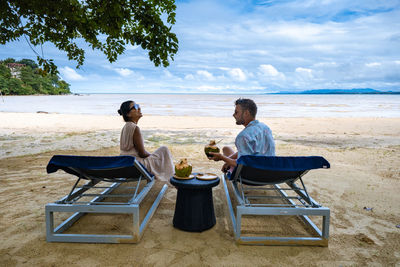 Rear view of woman sitting on lounge chair at beach against sky