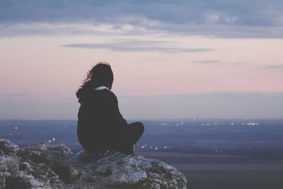 Rear view of woman sitting on rock against sky during sunset