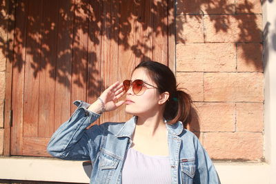 Portrait of young woman wearing sunglasses standing against wall