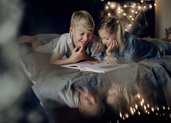 Smiling siblings reading book while lying on bed