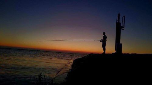 Silhouette men standing on cliff by sea against sky during sunset