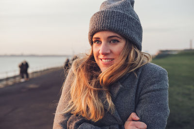 Cheerful young female in stylish knitted clothes and hat sitting on weathered wooden bench and enjoying sunny autumn day during travel in nature