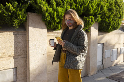 Young woman wearing face mask holding disposable cup and smart phone outdoors