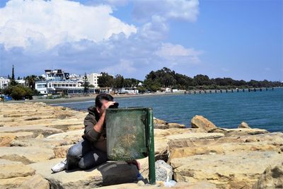 Full length of man looking at sea through binoculars by garbage bin on rocks against sky