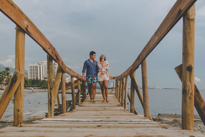 Full length of cheerful couple walking on pier at beach against sky