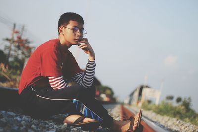 Tilt shot of teenage boy sitting on railroad tracks against sky