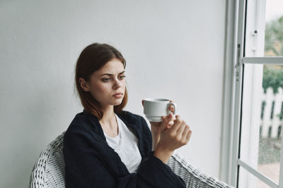 Young woman drinking coffee while sitting on window