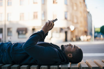 Side view of man lying on bench while using smart phone in city