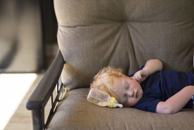 Cute baby boy sleeping by milk bottle on couch at home