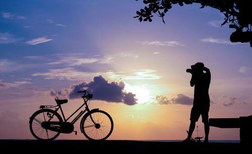 Silhouette man photographing bicycle against sky during sunset
