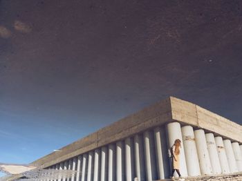 Woman walking by building against sky during sunny day