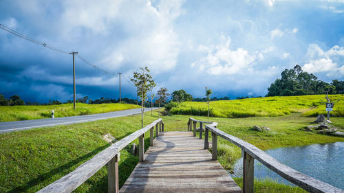 Boardwalk on grassy field by road against cloudy sky