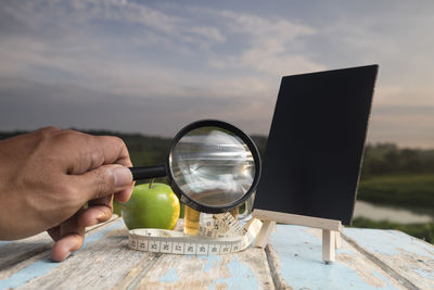 Cropped hand holding magnifying glass at table against sky