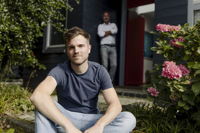 Adult son sitting in backyard while father leaning on house wall