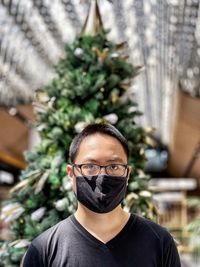 Portrait of young man wearing eyeglasses and face mask against christmas tree and skylight.