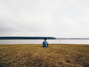 Rear view of man sitting at riverbank against sky