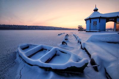 Snow covered building by lake against sky during sunset