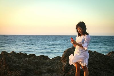 Young woman using mobile phone while standing at beach against clear sky during sunset