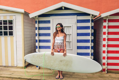 Young woman with surfboard standing against beach hut
