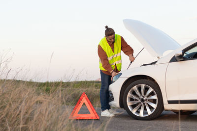 Young man on vest with red warning triangle sign off road trying to fix his broken car