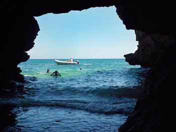 People swimming in sea seen through cave against clear sky