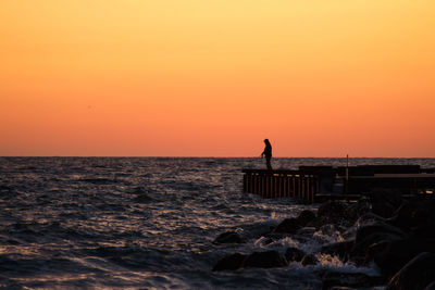 Silhouette man fishing on pier over sea against orange sky