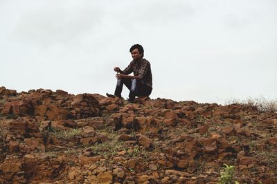 Young man sitting on rock against sky