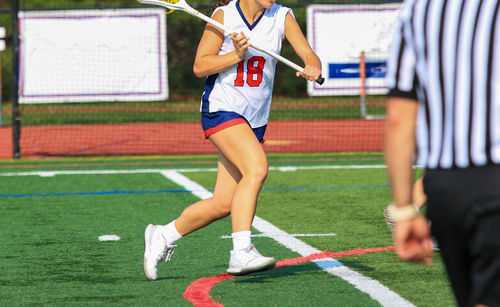 A female player in uniform runs with a lacrosse stick, focused and determined on the field.