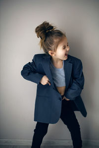 Funny portrait of fashionable girl child in large gray jacket stands against gray wall next  window 