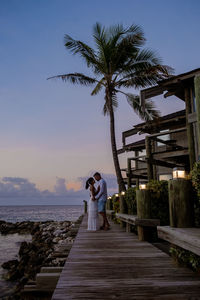 Couple standing by palm tree by sea against sky