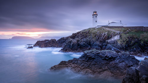 Lighthouse on rocks by sea against sky during sunset